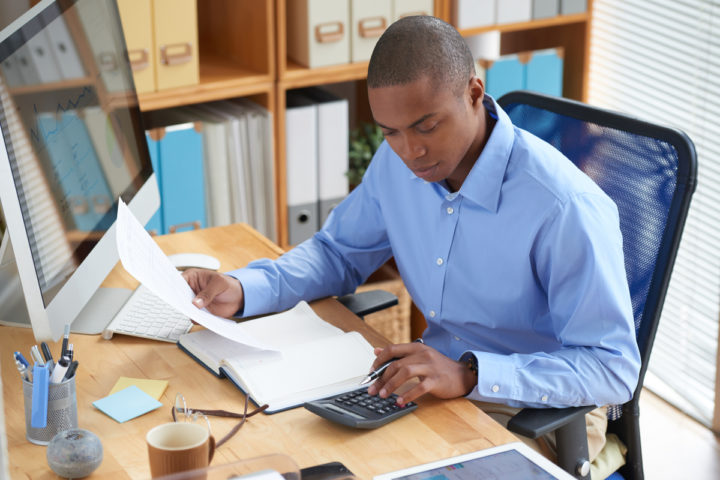 Homem negro de camiseta azul checando relatório em frente ao computador.