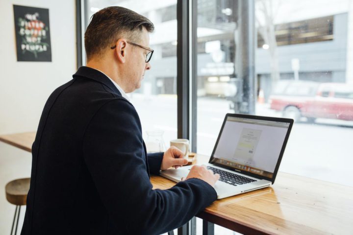 Homem caucasiano de meia idade navega na internet em um notebook apoiado sobre uma bancada de madeira e segura um copo de café.